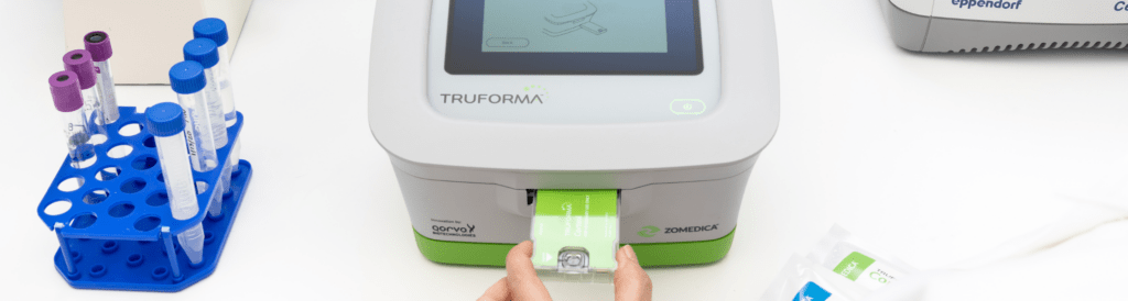 Hands inserting a test cartridge into the TRUFORMA point-of-care diagnostic device, with sample tubes and a lab analyzer in the background